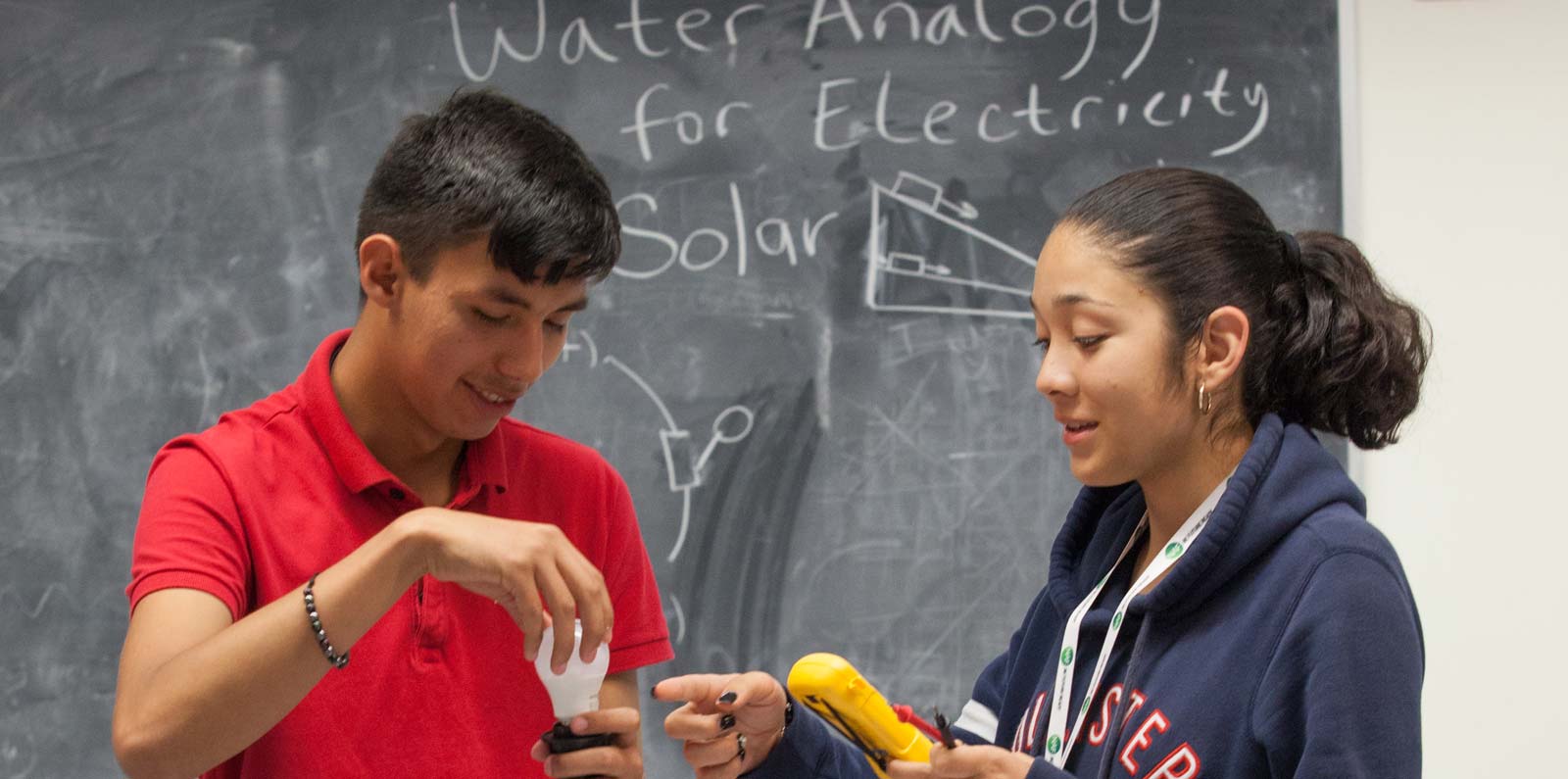 Teenaged students work on an experiment including a light bulb circuit and an volt meter with the words " Water Analogy for Electricity" on a chalkboard behind them.